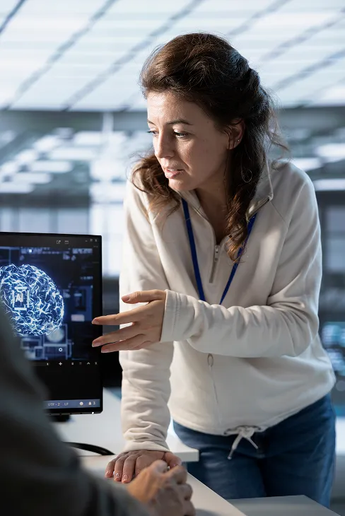 Woman pointing at a monitor showing an AI brain visualization while talking to a colleague